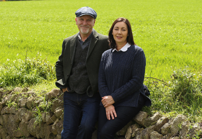 Mike and Carina Collins sitting on a stone wall in front of a green field in Ireland